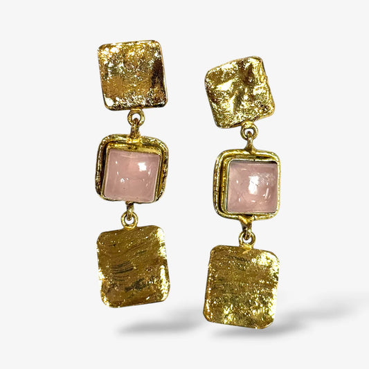 A pair of gold drop earrings featuring textured gold square plates, a central pale pink square stone, and a larger textured gold square at the bottom, shown against a light gray background.