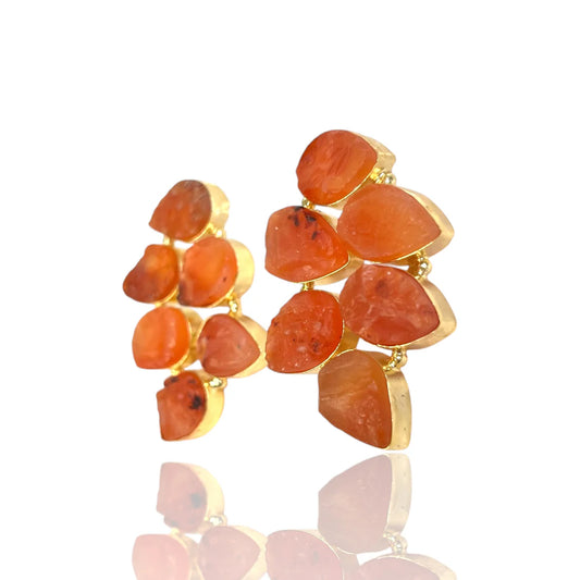 A pair of gold statement earrings featuring irregular, organic-shaped orange gemstones, displayed on a clean white background with a soft reflection below.