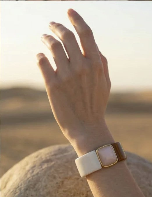 A close-up of a person's hand and wrist wearing a fashion bracelet with a dual-tone band, featuring a cream-colored acrylic section, a brown  section, and a gold-toned bezel with a pale ivory inlay.